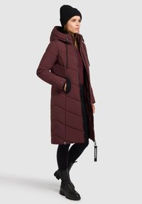 Burgunderfarbener Puffer-Mantel mit Kapuze, der ein Zickzackmuster, einen doppelten Reißverschluss und schwarze Akzente aufweist. Kombiniert mit schwarzen Leggings und Stiefeln.