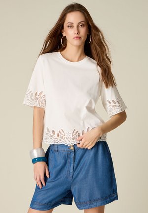 Ung kvinde med langt brunt hår iført hvid kortærmet top med blondekant og løst blå denimshorts, accessoriseret med store creoler og chunky armbånd.