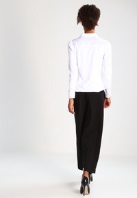Weißes Langarmhemd mit Kragen, kombiniert mit schwarzen, weit geschnittenen Hosen. Das Hemd hat geknöpfte Manschetten und die Hosen haben eine glatte Textur.