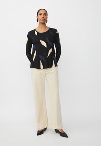 Donna che indossa un top nero a maniche lunghe con motivi astratti di foglie beige e blu navy, pantaloni larghi color crema, scarpe nere con punta e orecchini neri.