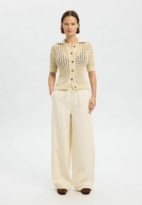 Cardigan en tricot beige avec un design à mailles ouvertes, manches courtes et fermeture à boutons. Associé à un pantalon ample de couleur claire.