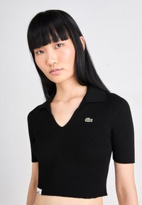 Polo shirt negru scurt, cu mâneci scurte, textură ribbed, design în formă de V și un mic logo verde Lacoste pe piept.