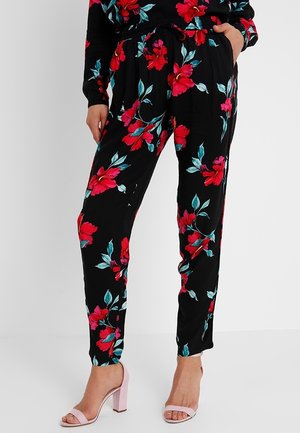 Žena v černých kalhotách s květinovým vzorem v červené a tyrkysové barvě, doplněných světle růžovými sandály s otevřenou špičkou a blokovým podpatkem.