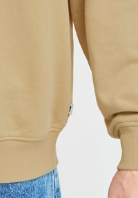 Beige sweatshirt met ribbels aan de manchetten en onderkant. Zachte textuur met een klein merklabel bij de zijnaad. Draagt met blauwe spijkerbroek.