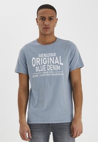 Lyseblå bomull T-skjorte med en grafisk trykk i hvitt med teksten "Genuine Original Blue Denim" og slagorddetaljer nedenfor.