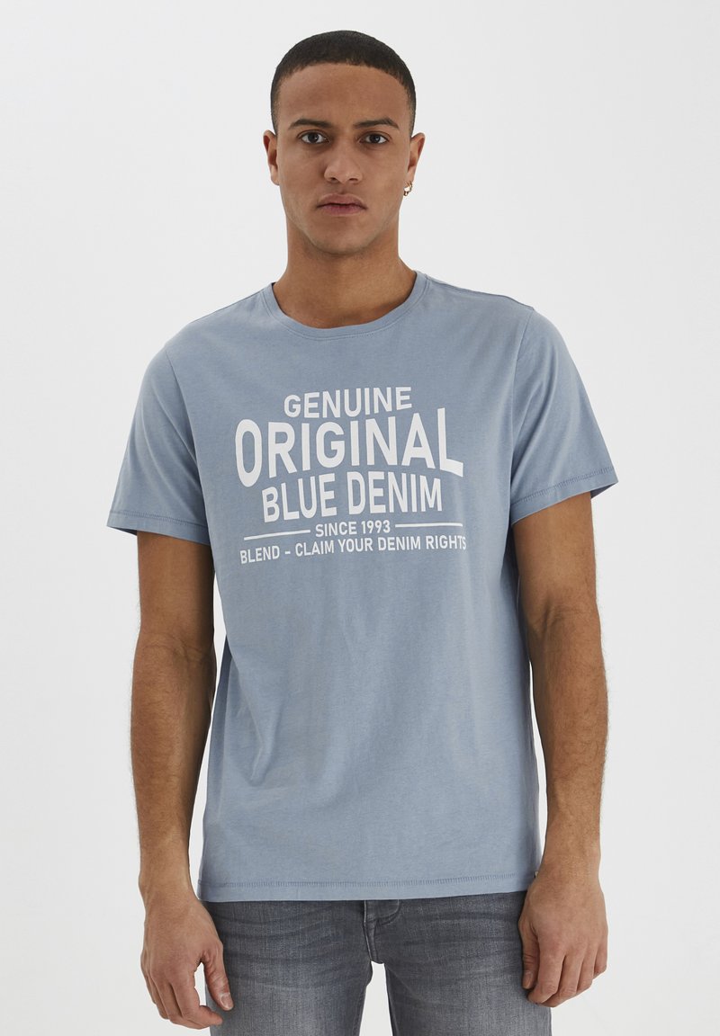 Lyseblå bomull T-skjorte med en grafisk trykk i hvitt med teksten "Genuine Original Blue Denim" og slagorddetaljer nedenfor.