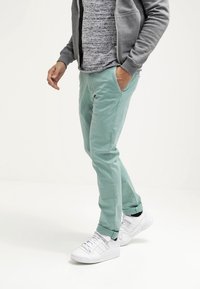 Mátově zelené chino kalhoty vyrobené z lehkého materiálu, s bočními kapsami a slim fit střihem. V kombinaci s bílými teniskami a šedou šusťákovou bundou na zip.