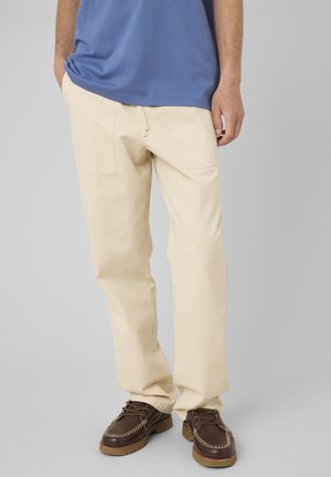 Homme portant un pantalon couleur crème, des chaussures marron à lacets et une chemise bleue, debout avec une main dans la poche sur un fond gris.