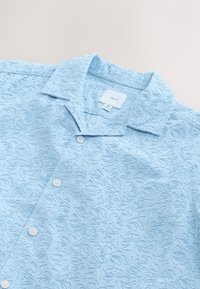 Chemise à manches courtes bleu clair présentant un motif floral en relief, fermeture par boutons et col classique. Étiquette de la marque visible à l'intérieur.