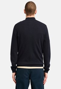 Dark navy knitted sweater with long sleeves, a round neckline, and a soft texture, worn over a light-colored shirt and navy pants.