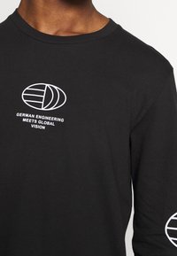 Czarna koszulka z długim rękawem, wykonana z bawełny, z białymi nadrukami na przodzie i rękawie, z napisem "NIEMIECKIE INŻYNIERIA SPOTYKA GLOBALNĄ WIZJĘ."