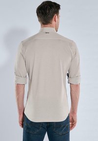 Mann in beigem Langarmhemd mit hochgerollten Ärmeln und blauen Jeans, von hinten vor einem schlichten Hintergrund gezeigt.