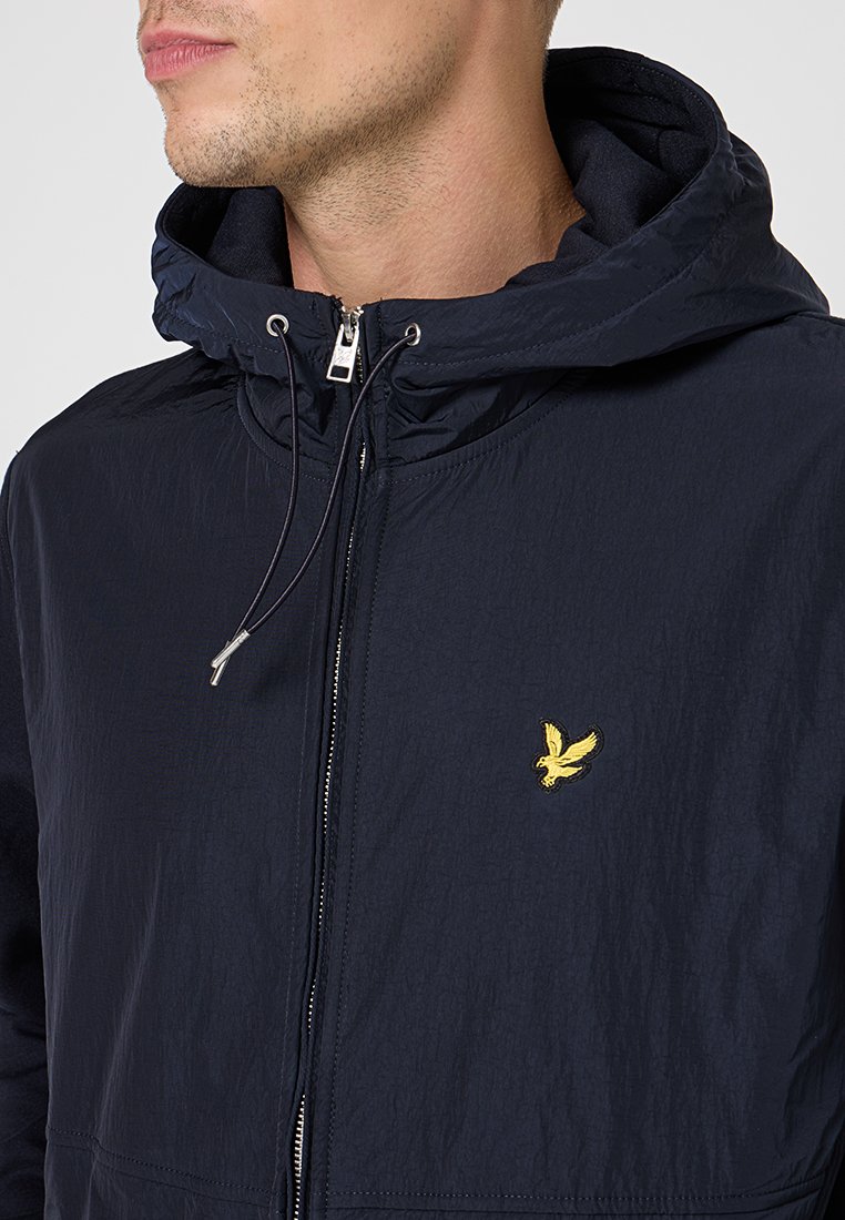 Veste coupe-vent navy avec une texture légère, une capuche, une fermeture éclair et un logo d'aigle jaune sur la poitrine. Design décontracté.