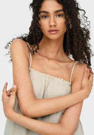 Giovane donna con capelli ricci scuri che indossa una canotta beige senza maniche e orecchini a cerchio dorati, con le braccia incrociate delicatamente sul petto.