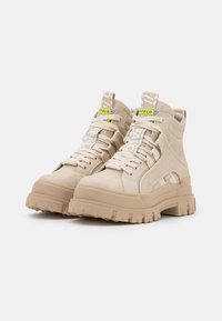 Bottines beige en daim, présentant une semelle épaisse, un design à lacets et des sangles de marque distinctives avec des accents en jaune et blanc contrastants.