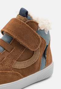 Chaussure pour tout-petits en suède marron avec des accents bleus, fermeture à bande Velcro, doublure en polaire en haut, et semelle en caoutchouc blanche.