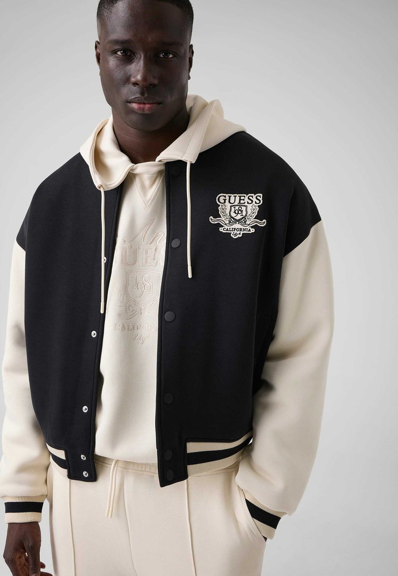 Homme portant une veste universitaire GUESS noire et crème sur un hoodie crème et un pantalon de survêtement, debout devant un fond clair uni.
