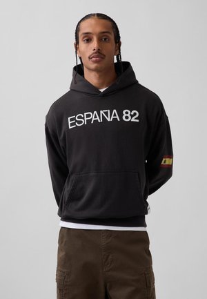 Bărbat cu părul împletit poartă un hanorac negru cu textul „ESPAÑA 82” și un mic patch cu steagul Spaniei pe mâneca stângă.
