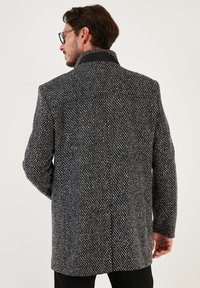 Abrigo texturizado en blanco y negro con un alto cuello, con un corte clásico y un sutil patrón de pata de gallo. Detalles de costura visibles en el cuello.