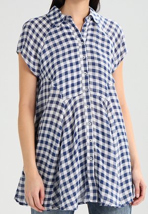 Niebiesko-biała tunika w kratę vichy z krótkim rękawem, zapinana na guziki, z kołnierzykiem, luźnego kroju, noszona na dżinsach.