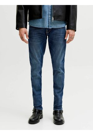 Hombre vestido con vaqueros ajustados azules, chaqueta de cuero negra, camisa de mezclilla y botines de cuero negros, posando frente a un fondo liso.