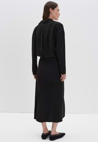 Femme se tenant de côté, portant un blouse noire à manches longues, une jupe midi noire et des chaussures plates noires avec des détails en œillets.