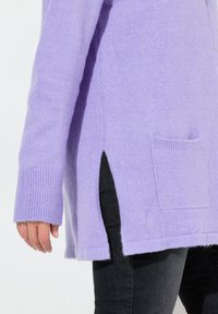 Cardigan in lana lilla con polsini a costine, fessure laterali e una tasca davanti. La texture sembra morbida e lavorata a maglia, indossato sopra pantaloni neri.