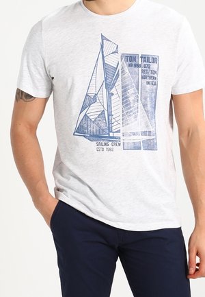 Man in een lichtgrijs T-shirt met een blauwe zeilboot afbeelding en tekst, gecombineerd met donkerblauwe broek, staand met één hand in zijn zak.