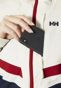 Black smartphone case being placed into a zipper pocket of a cream jacket with red and navy stripes, featuring a logo on the chest.