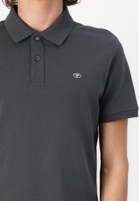 Mørkegrå polo skjorte med strukturert stoff, med en tre-knappers placket og en liten logo på venstre bryst. Korte ermer.