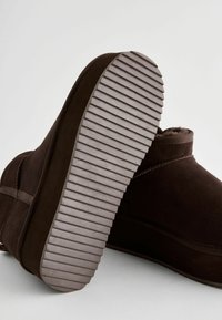 Bottines en daim marron avec un intérieur doux, dotées d'une semelle en caoutchouc gris texturé et d'une forme arrondie. Des détails de couture subtils sont visibles.