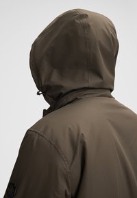 Olivegrüne Kapuzenjacke mit strukturiertem Finish, ausgestattet mit einem schwarzen Reißverschluss und einem Logodetail auf der Schulter. Verstellbares Kapuzendesign.