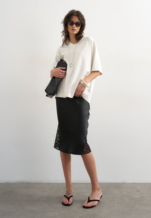 Femme portant un t-shirt blanc oversize, jupe noire bordée de dentelle, sandales noires, lunettes de soleil, tenant une grande pochette noire contre un mur uni.