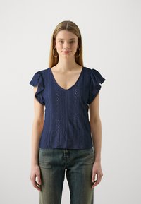 Marineblå topp med korte volangermer, V-utringning, teksturert mønster med hullmønster, kombinert med falmede blå jeans.