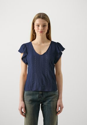 Haut bleu marine avec de courtes manches volantes, décolleté en V, motif texturé avec des détails en œillet, associé à un jean bleu délavé.