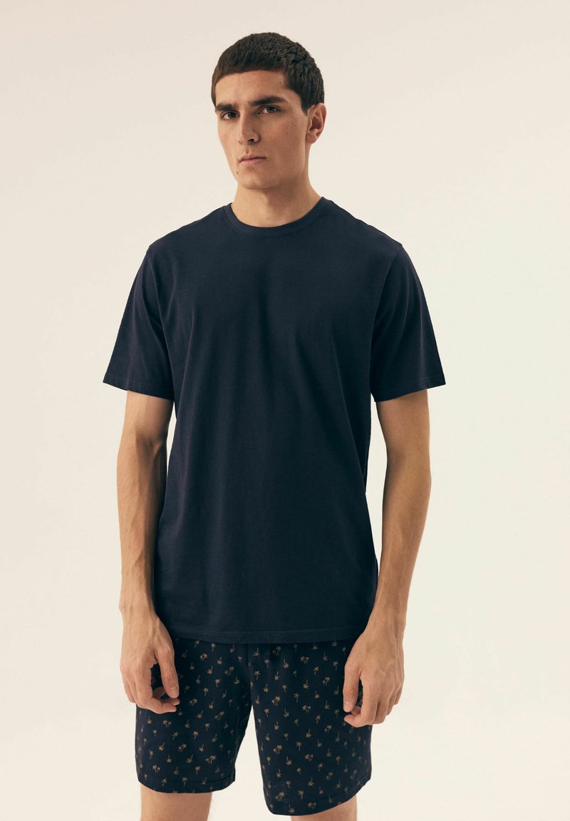 Granatowa bawełniana koszulka z krótkim rękawem w połączeniu z ciemnymi spodniami z wzorem, na których znajdują się małe motywy palm, wyeksponowana na neutralnym tle.