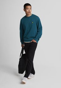 Sudadera verde azulado con un pequeño logo amarillo, combinada con pantalones negros y zapatillas grises. La persona lleva una bolsa de tela negra.