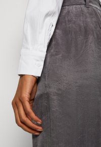 Main reposant contre une jupe texturée grise, portant une chemise blanche à manches longues avec un poignet boutonné.