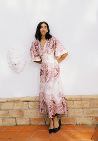 Femme en longue robe blanche avec des motifs roses et des talons noirs se tient contre un mur blanc avec une tête de lion décorative.