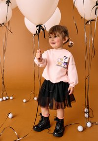 Rosa sweatshirt med tecknad design, svart tyllkjol och svarta snörstövlar. Håller i en ballong, omgiven av ballonger och dekorativa element.