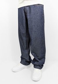 Denim jeans in dark blue with a loose fit, featuring visible stitching and a straight leg. Paired with white sneakers.
