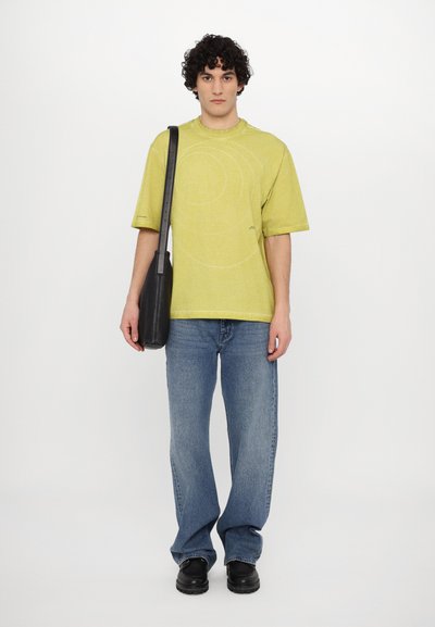 Lime green oversized t-shirt with circular pattern, paired with blue wide-leg jeans and black shoulder bag. Model wearing black shoes.