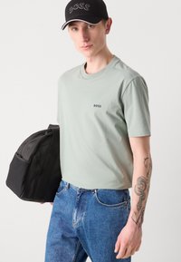 Lys grønn bomull T-skjorte med "BOSS" logo, sammen med blå denimjeans. Svart stoffveske holdt i den ene hånden.