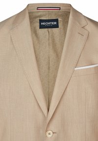 Blazer beige avec une texture lisse, revers crantés, fermeture à un bouton, et un mouchoir de poche blanc. Doublé d'un tissu marron clair à motif.