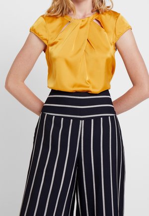 Kobieta w musztardowożółtej bluzce z krótkim rękawem i plisami oraz czarnych, szerokich spodni z wysokim stanem i białymi, pionowymi paskami.