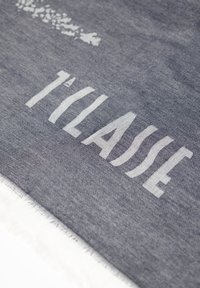 Tessuto grigio con testo bianco che recita "TILASSE," con una finitura morbida e testurizzata e bordi frangiati. Motivi sottili nel materiale.