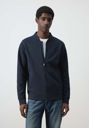 Joven con una chaqueta azul marino con cremallera sobre una camisa blanca y vaqueros azules, de pie frente a un fondo claro y liso.