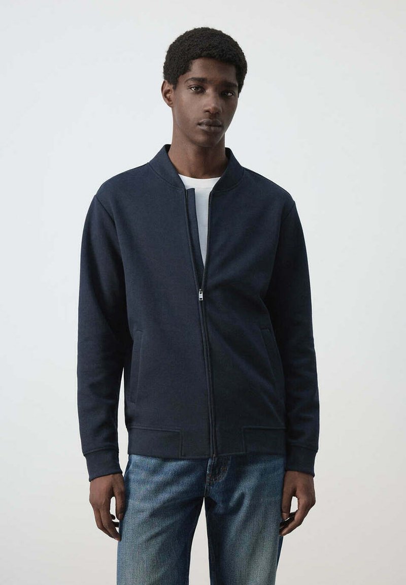 Jeune homme portant une veste zippée bleu marine par-dessus une chemise blanche et un jean bleu, debout devant un fond clair uni.