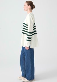 Pulover alb cu dungi orizontale verzi, purtat cu blugi cu talie înaltă și picioare largi de culoare albastră și adidași albi. Textură netedă și croială casual.