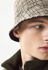 Brown and black checked bucket hat, with a wide brim and soft texture, paired with a dark green zip-up jacket. Close-up on face.
